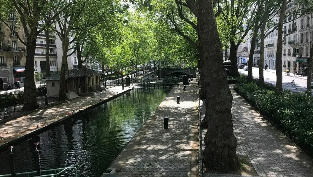 Le canal Saint-Martin (Paris-75010), un lieu serein bordé d'arbres et de sentiers pavés, flanqué de bâtiments de la ville sous un ciel clair.