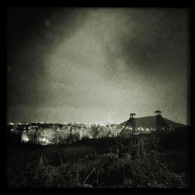 Photo en noir et blanc d'un paysage urbain la nuit avec des bâtiments et des structures industrielles contre un ciel nuageux. Auteur : Stéphanie Debaille.